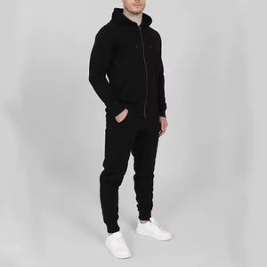 A man wearing a black zip-up hoodie, black jogger pants, and white sneakers, standing against a light gray studio background.