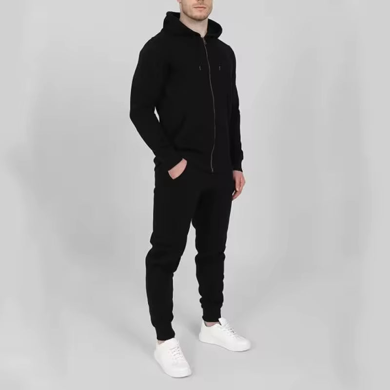 A man wearing a black zip-up hoodie, black jogger pants, and white sneakers, standing against a light gray studio background.